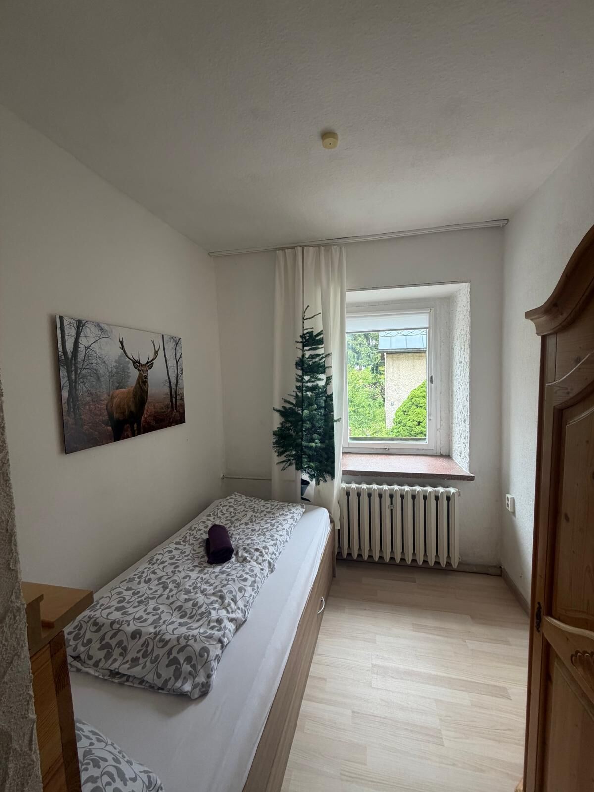 Un dormitorio pequeño con cama, radiador, ventana con vistas y un cuadro en la pared.