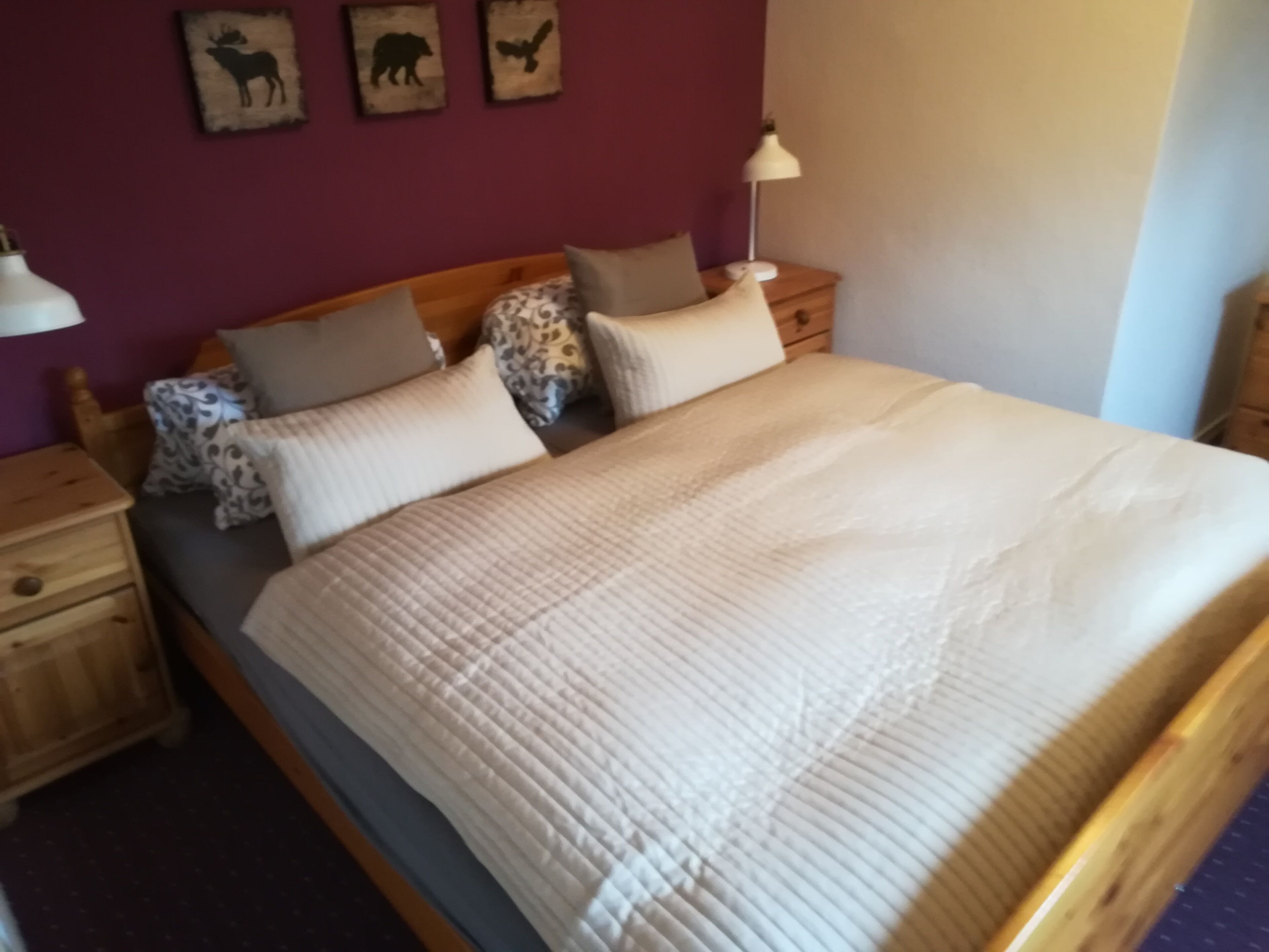 Una cama con cubrecolcha blanco acolchado, cabecera de madera y varias almohadas. Se observan obras de arte en la pared sobre la cama y una lámpara de noche en la mesita.