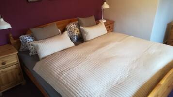 Una cama con cubrecolcha blanco acolchado, cabecera de madera y varias almohadas. Se observan obras de arte en la pared sobre la cama y una lámpara de noche en la mesita.