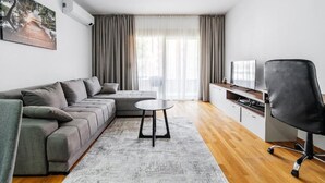 Una sala de estar moderna con un sofá seccional gris, una mesita redonda pequeña, una silla de oficina negra, un televisor de pantalla plana y un ventanal con cortinas.
