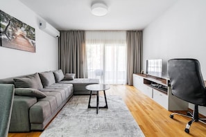 Ein modernes Wohnzimmer mit einem grauen Ecksofa, einem kleinen runden Tisch, einem schwarzen Bürostuhl, einem Flachbildfernseher und einem großen Fenster mit Vorhängen.
