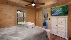 Ein Schlafzimmer mit Bett, Deckenventilator, Fernseher, Kleiderschrank und einem Fenster mit Blick auf Bäume.