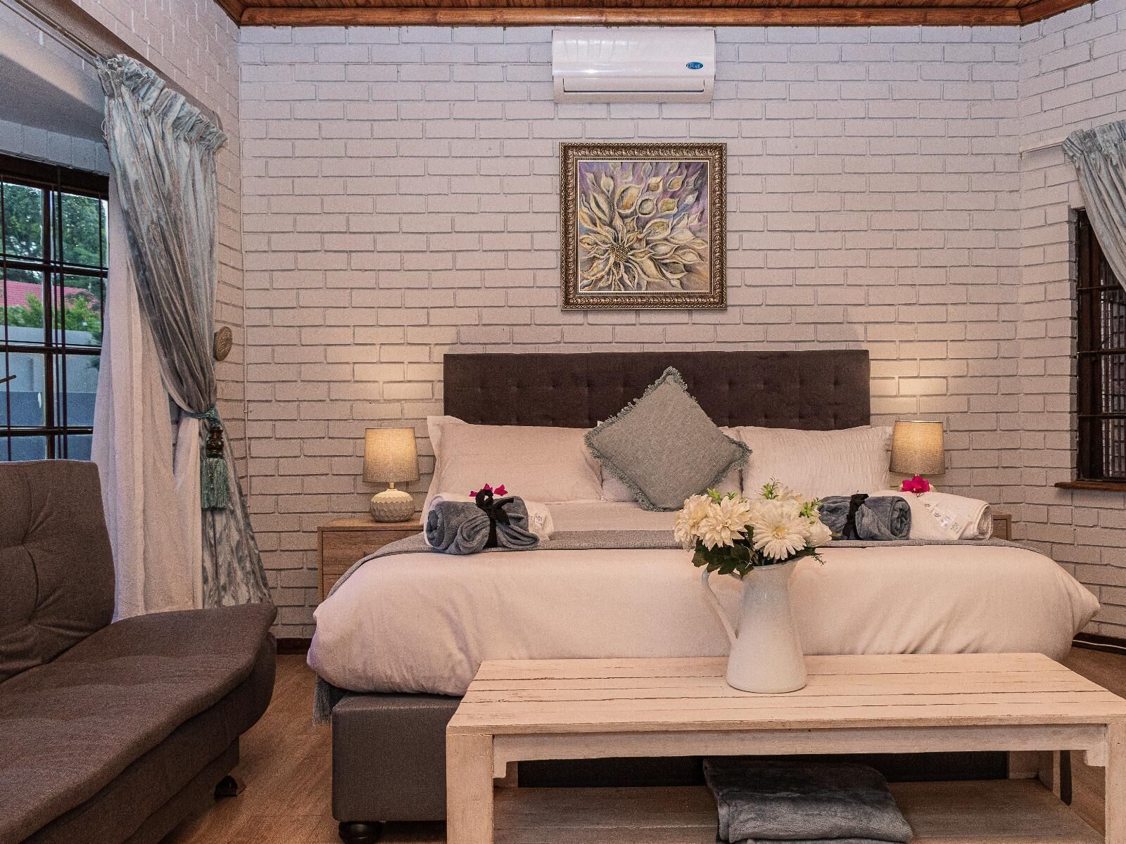 Un dormitorio con pared de ladrillo, una cama con almohadas, una mesita de noche con lámpara, una ventana con cortinas y un jarrón con flores sobre una mesa.