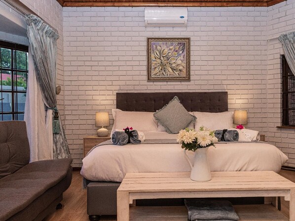 A bedroom with a brick wall, a bed with pillows, a nightstand with a lamp, a window with curtains, and a vase with flowers on a table. - Mereke Manor (Centurion)
