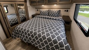 A bedroom with a patterned bedspread, a mirror, a window with a view, and built-in cabinets. - The TajMyHaul, Camper in Waterside at Blue ridge! (Morganton)