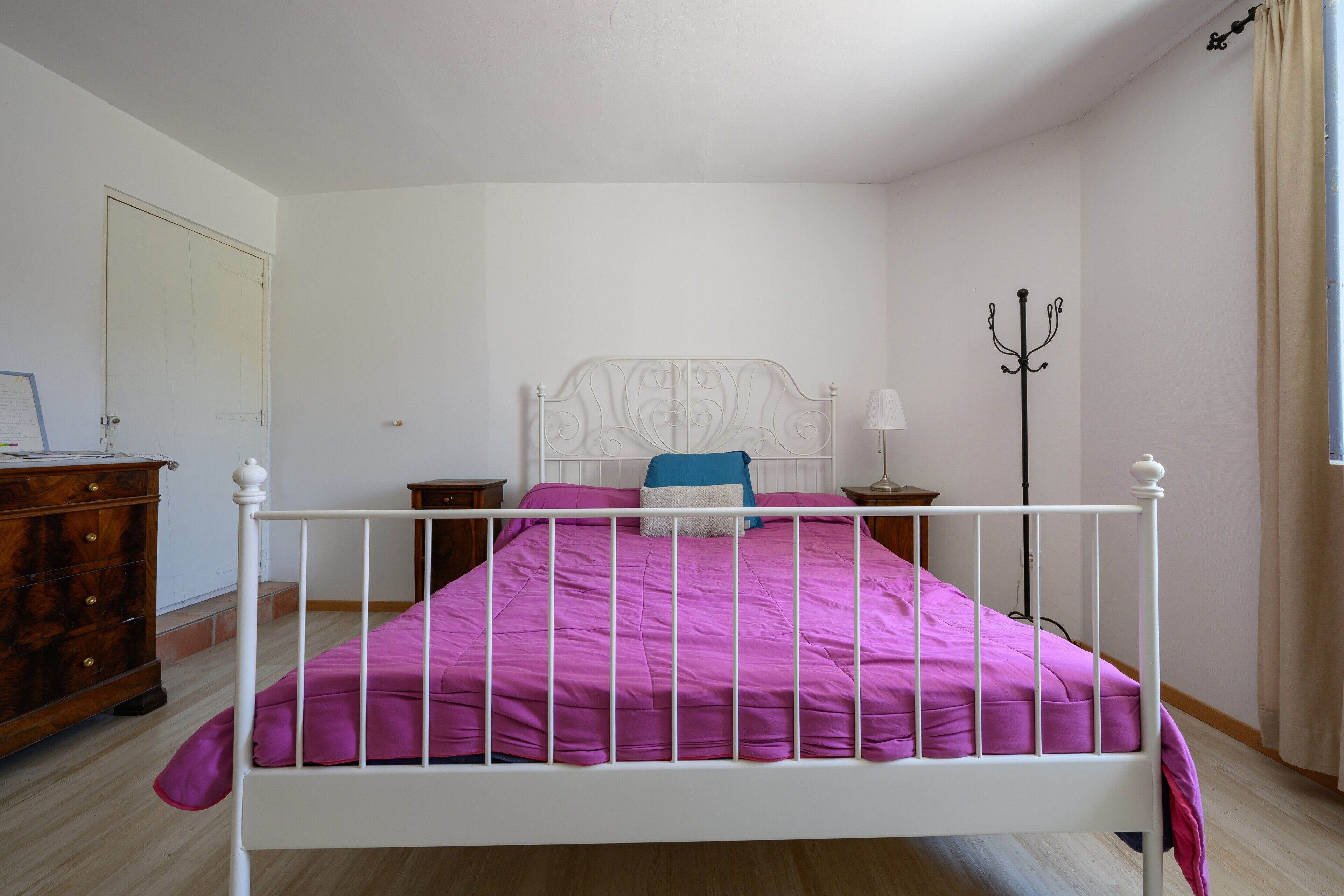 Una camera da letto con un letto in ferro bianco, una coperta viola, un comò in legno e una lampada da terra.