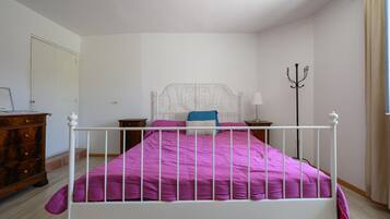 Una camera da letto con un letto in ferro bianco, una coperta viola, un comò in legno e una lampada da terra.