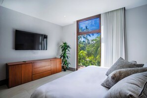 Ein Schlafzimmer mit Bett, Fernseher, Pflanze und Fenster mit Blick auf Grünflächen.