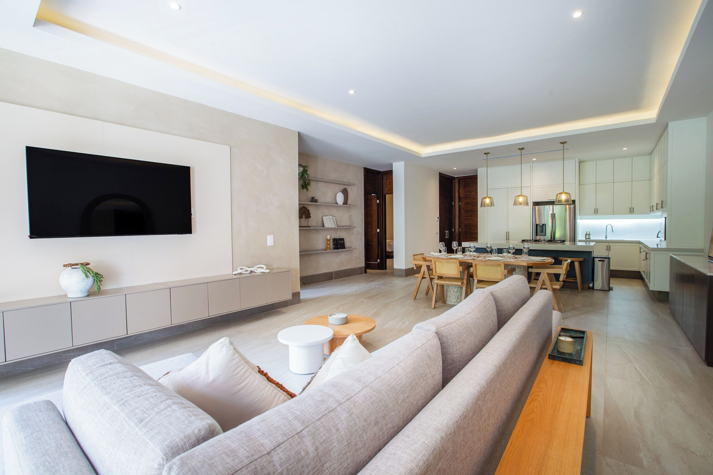 Ein modernes Wohnzimmer mit großem Flachbildfernseher, einer Couch, einem Essbereich mit Tisch und Stühlen sowie einer Küche mit Schränken und einem Kühlschrank.