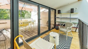 Et moderne værelse med en skydedør til en udendørs terrasse, en hvid fletstol, et lille bord og en ventilator på gulvet.