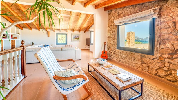 A spacious living area with a stone wall, a wooden ceiling, a large window with a view, a sofa, a chair, and a coffee table. - Finca in front of the sea, Can Cuarassa, Pollensa. (Pollensa)
