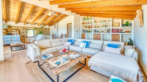 A spacious living room with a high ceiling, exposed wooden beams, a large sectional sofa, a coffee table, and a bookshelf filled with books and decorative items. - Finca in front of the sea, Can Cuarassa, Pollensa. (Pollensa)
