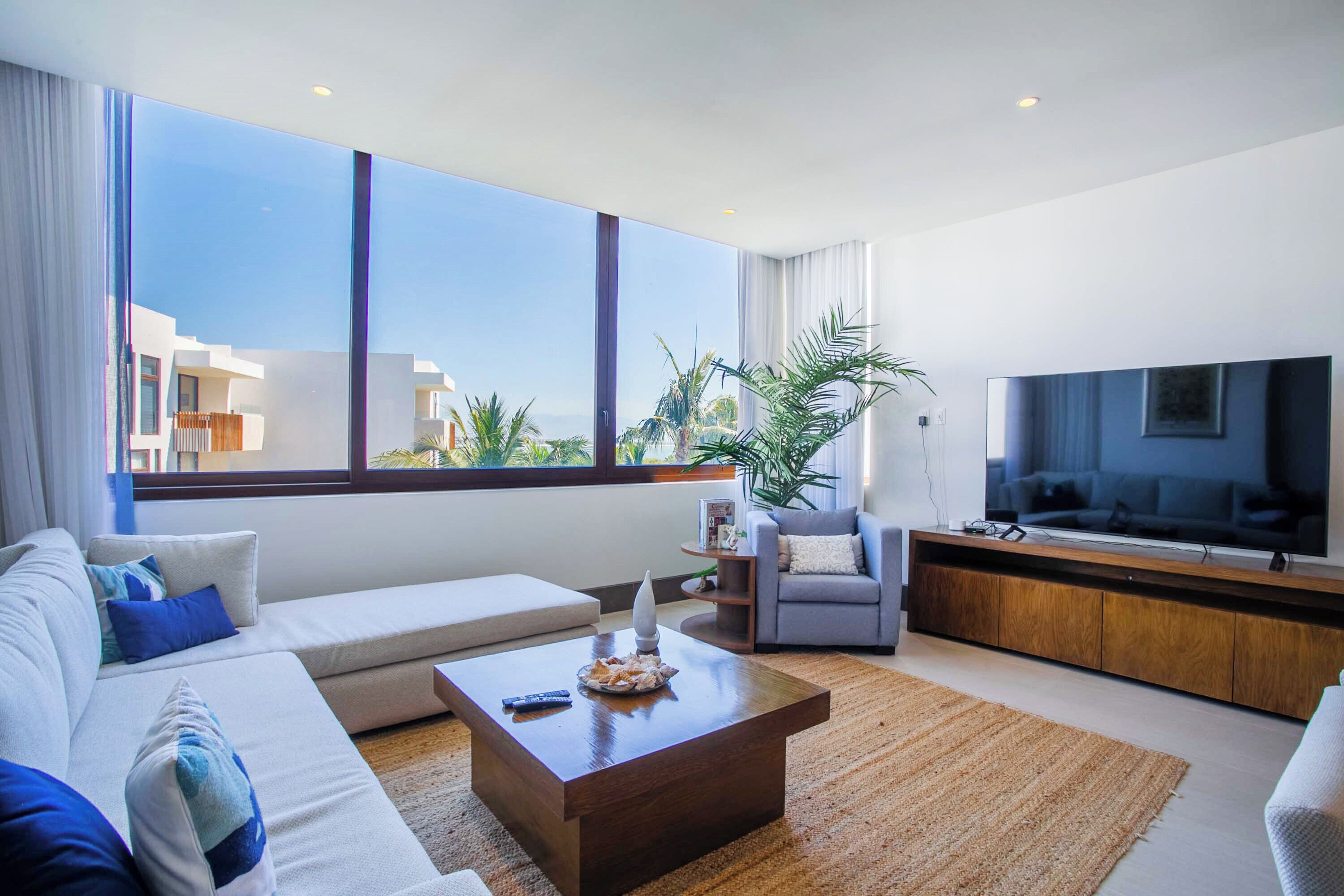 Uma sala de estar moderna com sofá, poltrona, mesa de centro, TV e grandes janelas.