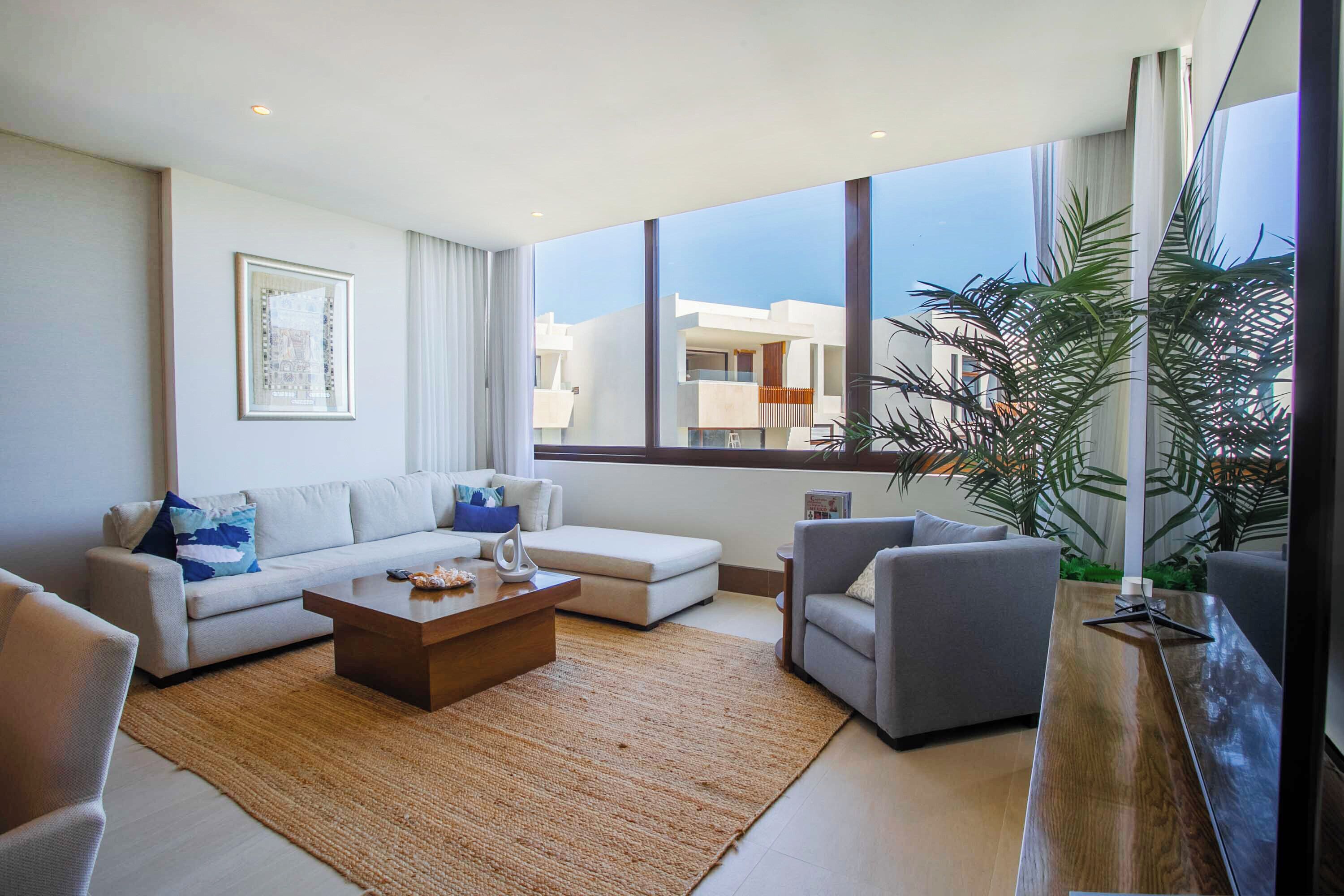 A modern living room with a sofa, armchair, coffee table, and large windows offering a view of buildings.