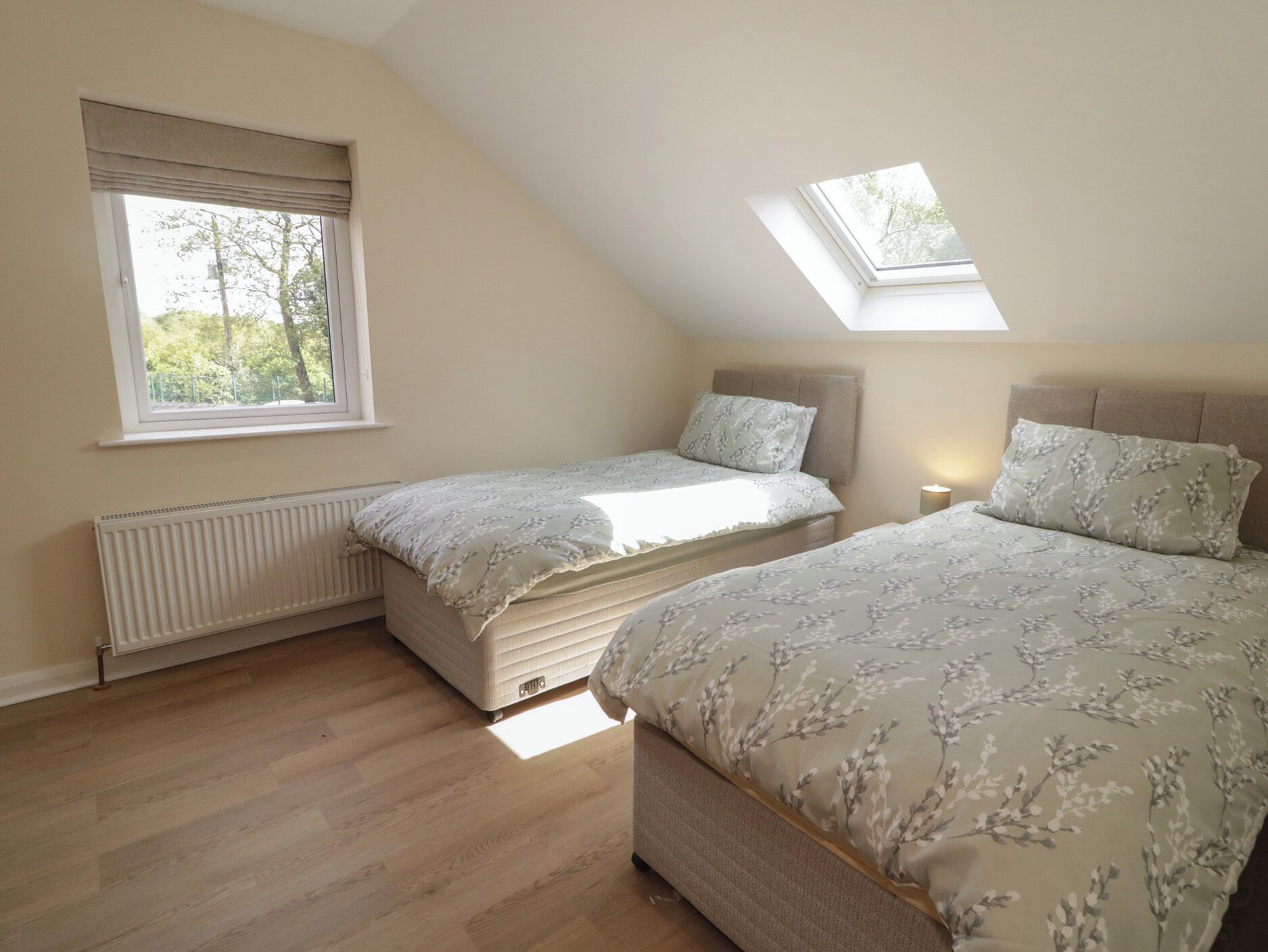 A bedroom with two beds, a skylight, a radiator, and a window with a view of trees.