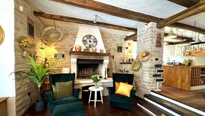 Een gezellige woonkamer met een open haard, twee groene fauteuils, een kleine tafel met een vaas, en een keuken in de achtergrond.