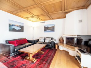 Ein Wohnzimmer mit Holzbalkendecke, Kamin, Fernseher, einer Couch mit roten Kissen und zwei an Wänden befestigten Bildern.