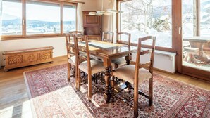 Uma sala de jantar com mesa e cadeiras de madeira, grandes janelas com vista para a neve e um tapete tradicional.