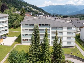 Ein mehrstöckiges Wohngebäude mit Balkonen, umgeben von Grünflächen und Bergen im Hintergrund.
