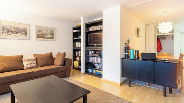 Ein modernes Wohnzimmer mit einer Couch, einem Couchtisch, einem Bücherregal, einem Fernseher und einem Schreibtisch mit Computer.
