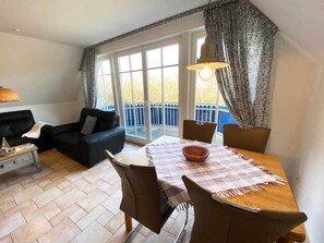 Ein Wohnzimmer mit einer Couch, einem Esstisch mit Stühlen, einer Lampe und Blick auf einen blauen Zaun durch große Fenster.