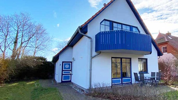 Une maison à deux étages avec un balcon, une porte dotée d’un cadre bleu et un petit espace extérieur aménagé pour s’asseoir.