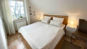 Une chambre à coucher avec un lit, des tables de chevet, une chaise, une fenêtre avec des rideaux et un radiateur.