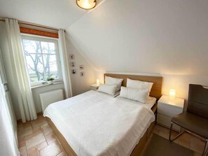 Ein Schlafzimmer mit einem Bett, Nachttischen, einem Sessel, einem Fenster mit Vorhängen und einem Heizkörper.