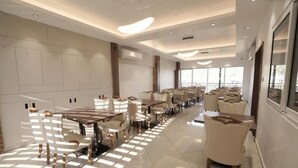 Een ruime hotel eetzaal met meerdere tafels en stoelen, grote ramen en een modern plafond.