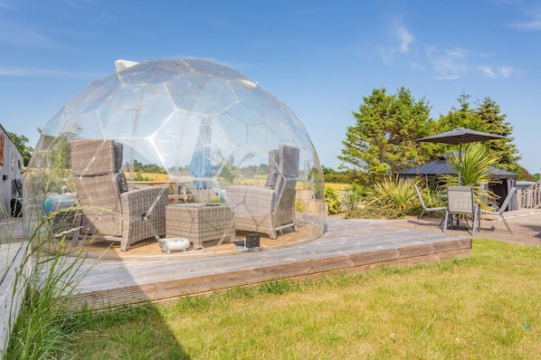 Una estructura de cúpula transparente con muebles de mimbre en su interior, rodeada por una terraza de madera y asientos exteriores.