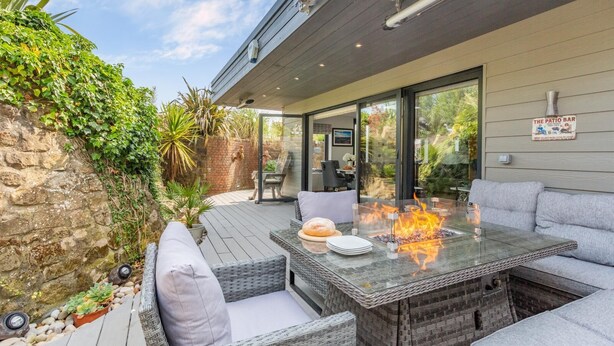 An outdoor patio with a glass-top table, fire feature, wicker furniture, and a stone wall. - Luxury Jurassic Spa Retreat | Private Swim Spa, Hot Tub & Gated Grounds (co durham)