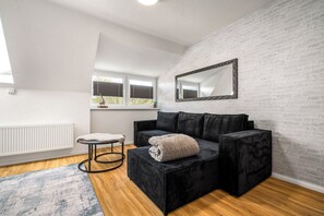 Een moderne kamer met een zwarte bank, een klein rond tafeltje, een witte radiator en een raam met zonwering.