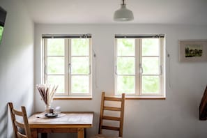 A room with two windows, a wooden table, a chair, a vase with dried plants, and a hanging light fixture.