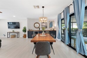 A modern dining area with a wooden table, chairs, a chandelier, and a large window with curtains. - 🏖️Sunshine Dock House | On Canal w/ Dock | Private Pool | Games 🏖️ (Fort Myers Beach)