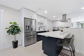 A modern kitchen with white cabinets, stainless steel appliances, a central island, and bar stools. - 🏖️Sunshine Dock House | On Canal w/ Dock | Private Pool | Games 🏖️ (Fort Myers Beach)