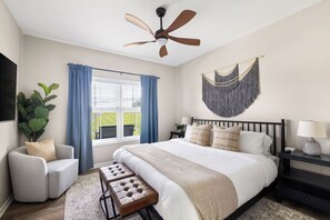 Ein Schlafzimmer mit Bett, Deckenventilator, Fenster mit Vorhängen, Pflanze, Bank und Wandleuchte.