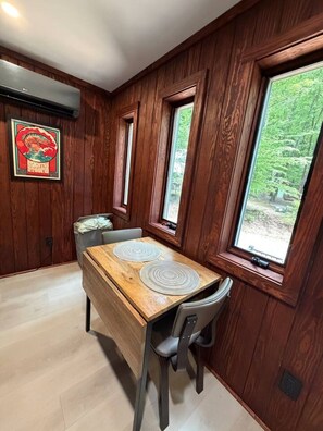A dining area with a wooden table and chairs, large windows with a view of greenery, and a wall-mounted air conditioning unit. - Ridgeline Treehouse at Cherryfield Luxury Camping (Brevard)