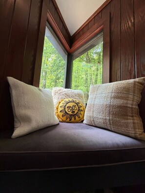 A window seat with three pillows, one with a sun design, and a view of a forest. - Ridgeline Treehouse at Cherryfield Luxury Camping (Brevard)