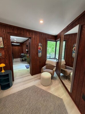 A room with wooden paneling, a large mirror, a beige armchair, a small ottoman, and a lamp. - Blueghost Treehouse at Cherryfield Luxury Camping (Brevard)