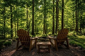 Dos sillas Adirondack de madera mirando hacia un hogar de piedra en un entorno boscoso.