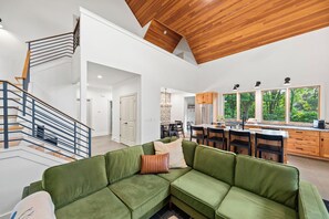 Ein modernes Wohnzimmer mit einem grünen Modulsessel, einer Holztreppe und einer Küche mit Holzausstattung und großen Fenstern.