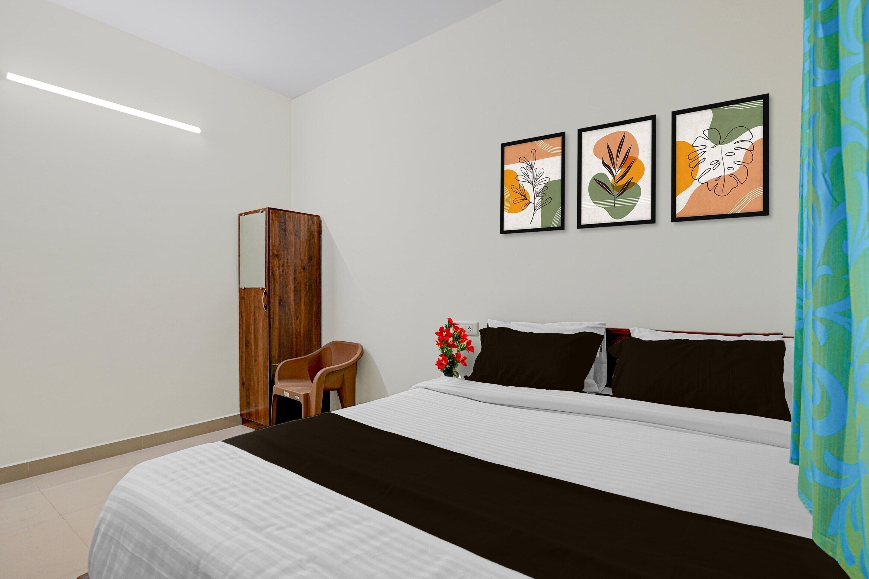 A hotel room with a bed, a wooden wardrobe, a chair, and three framed artworks on the wall.