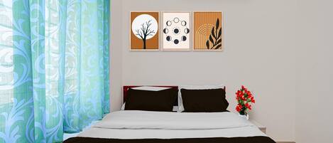 A hotel room with a bed, air conditioning unit, and three framed artworks on the wall.