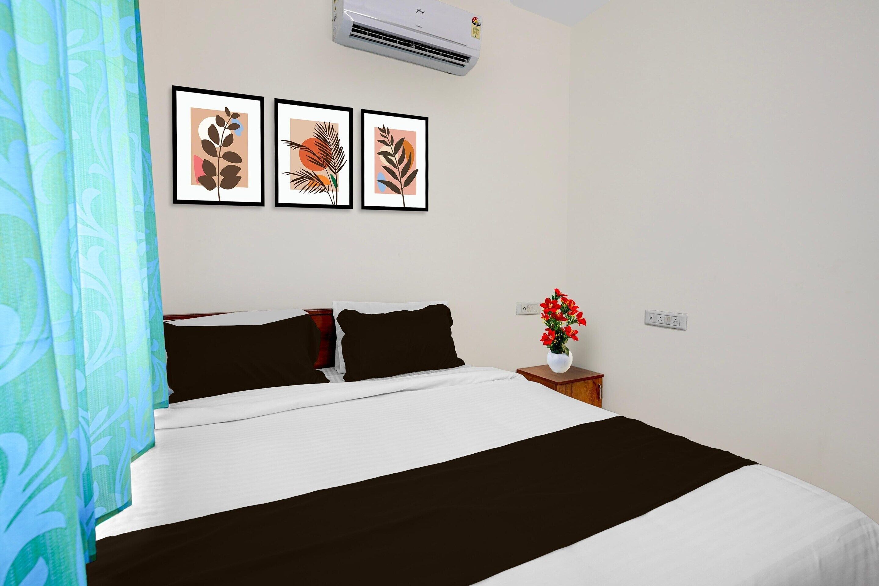 A hotel room with a bed, a nightstand with a vase of flowers, and three framed leaf artworks on the wall.