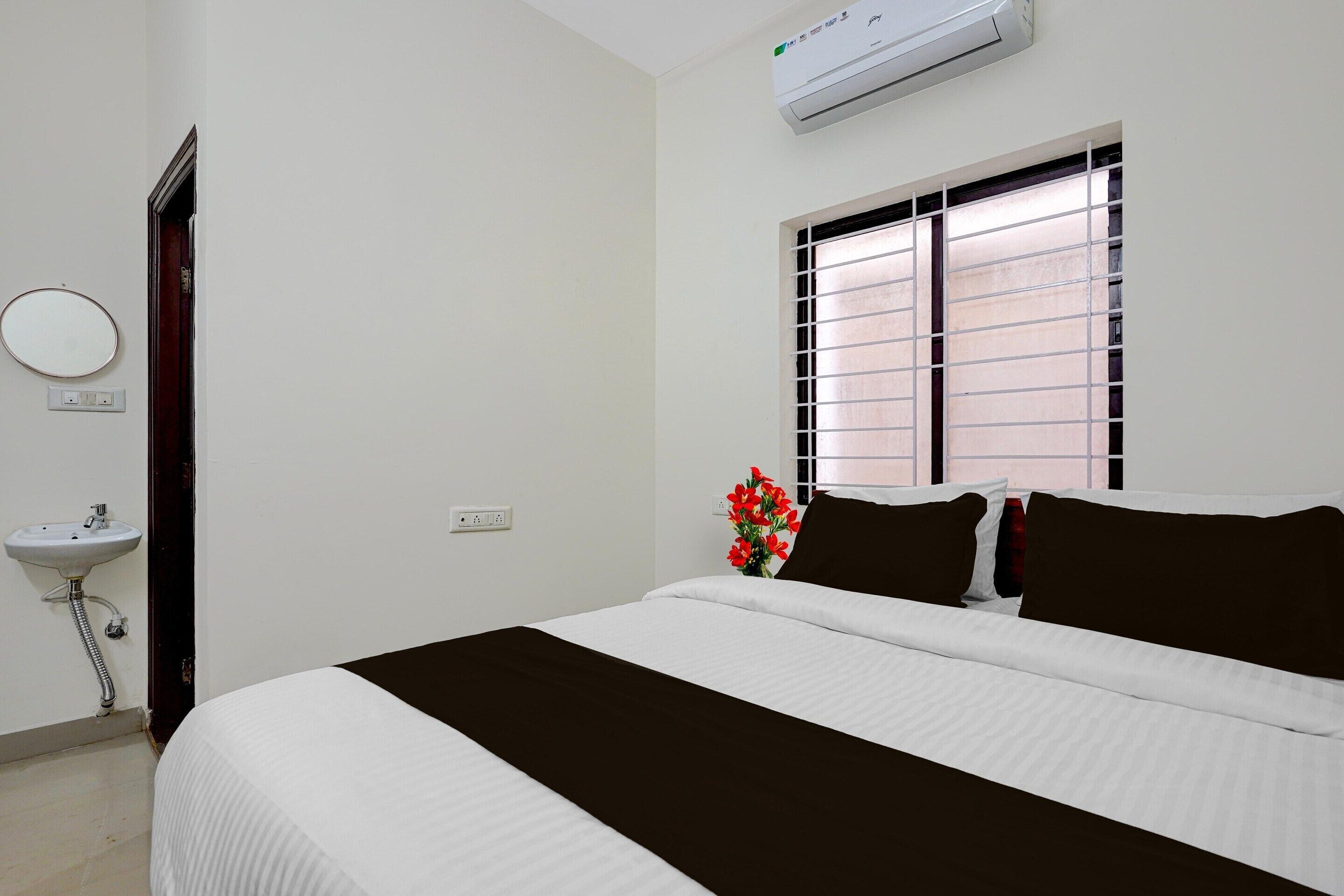 A hotel room with a bed, a window with blinds, a wall-mounted air conditioner, a bedside table, and a wall-mounted mirror.