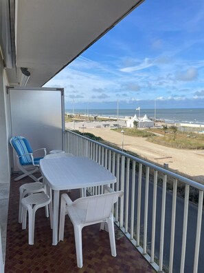 Un balcón con mesa y sillas blancas, vista a la playa y cielo despejado.