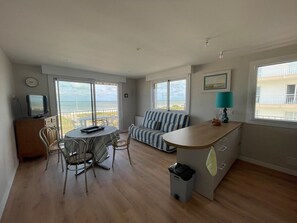 Quarto com mesa de jantar, cadeiras, sofá, pequena cozinha e vista para o oceano.