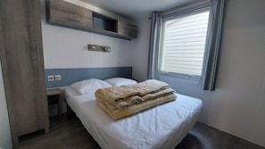 Een eenpersoonsbed met een witte matras en een beige deken, een nachtkastje, een houten plank boven het bed, een raam met zonwering, en een stopcontact op de muur.