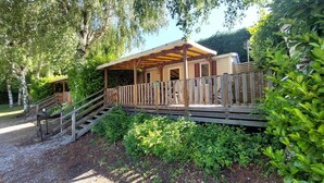 A wooden deck with a covered area, a staircase, and a building with red shutters. - Confort 2 Mobile Home with Mountain View, Wi-Fi & Air Conditioning (Saint-Laurent-en-Beaumont)