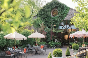 Un patio al aire libre con mesas y sillas, rodeado de abundante vegetación y un edificio de piedra con un ventanal circular.